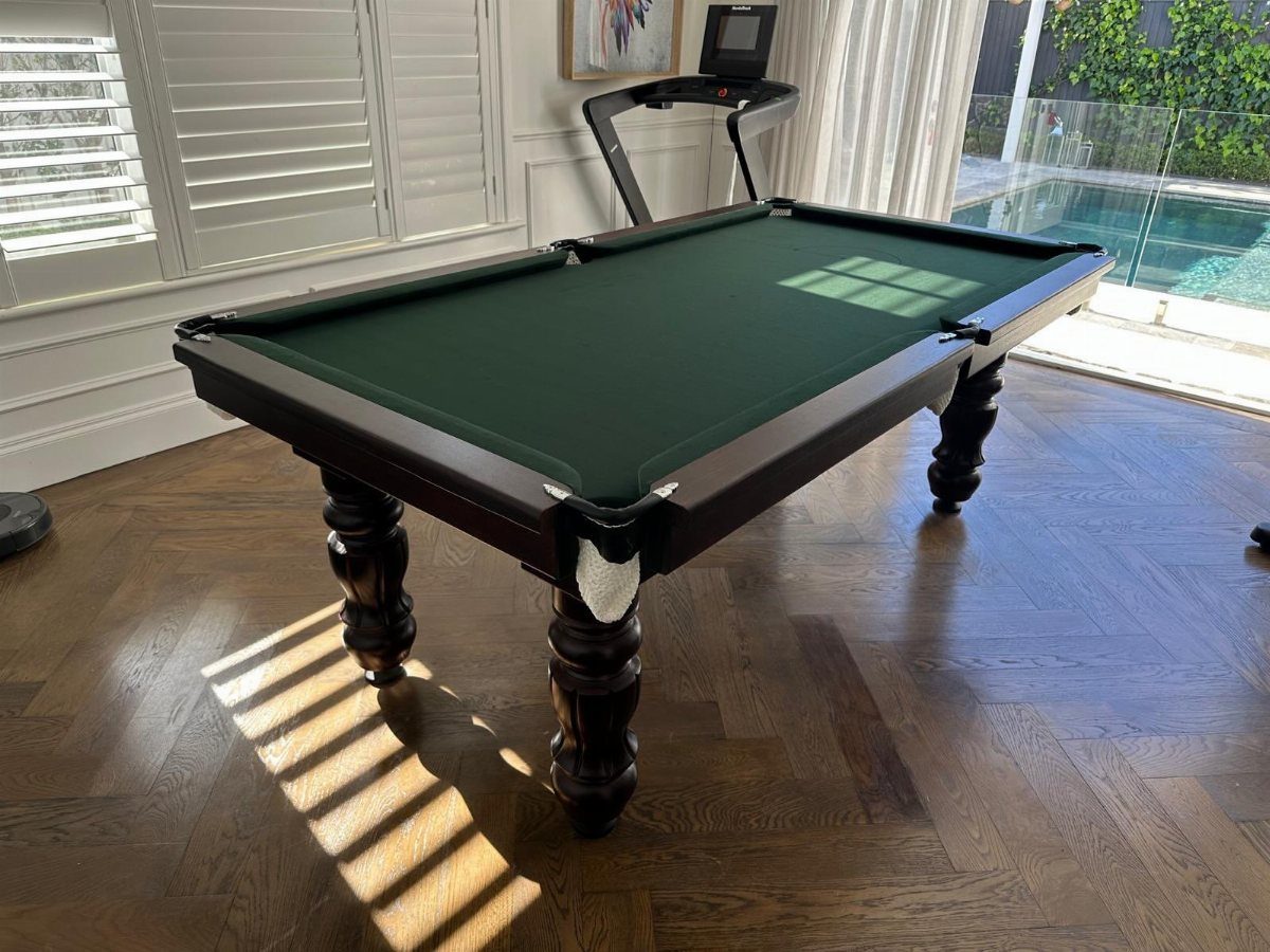 Dark wood pool table with green felt in a sunlit room with hardwood floors.