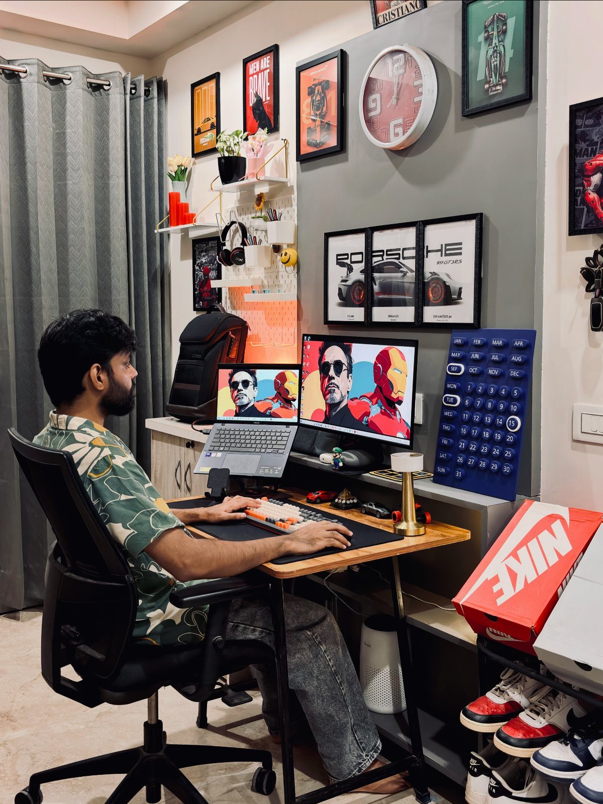Man working at a wooden desk with dual monitors and wall-mounted floating shelves.