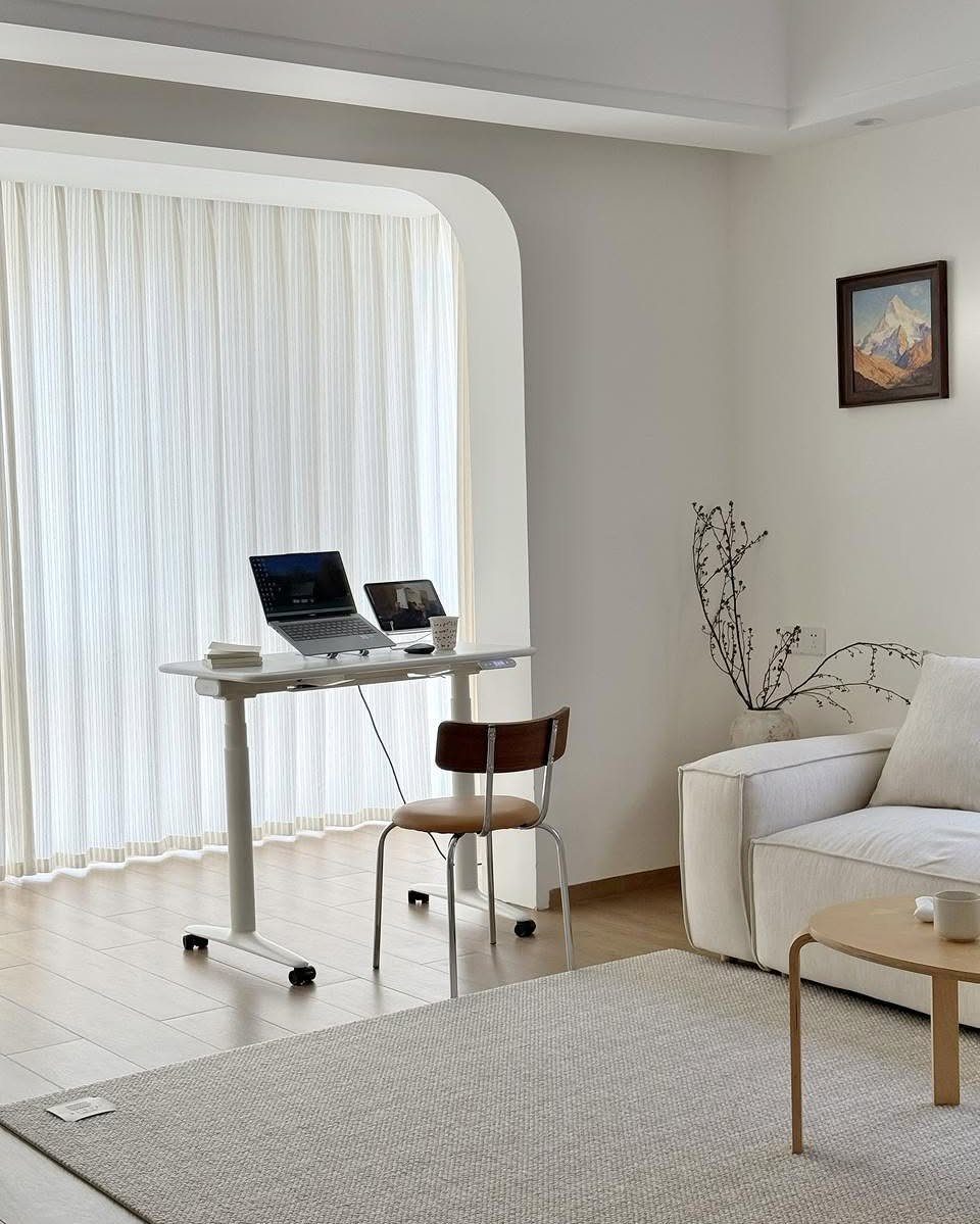 White adjustable desk with laptop and chair in a bright, modern living room.
