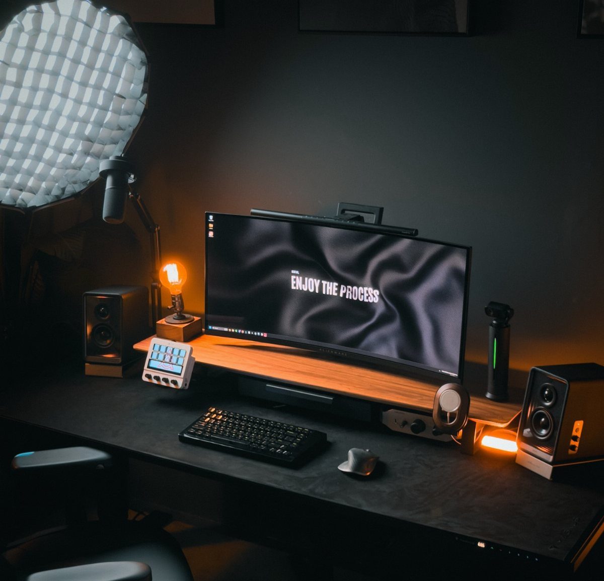 Black desk with wooden monitor riser, wide screen, and speakers in dark room