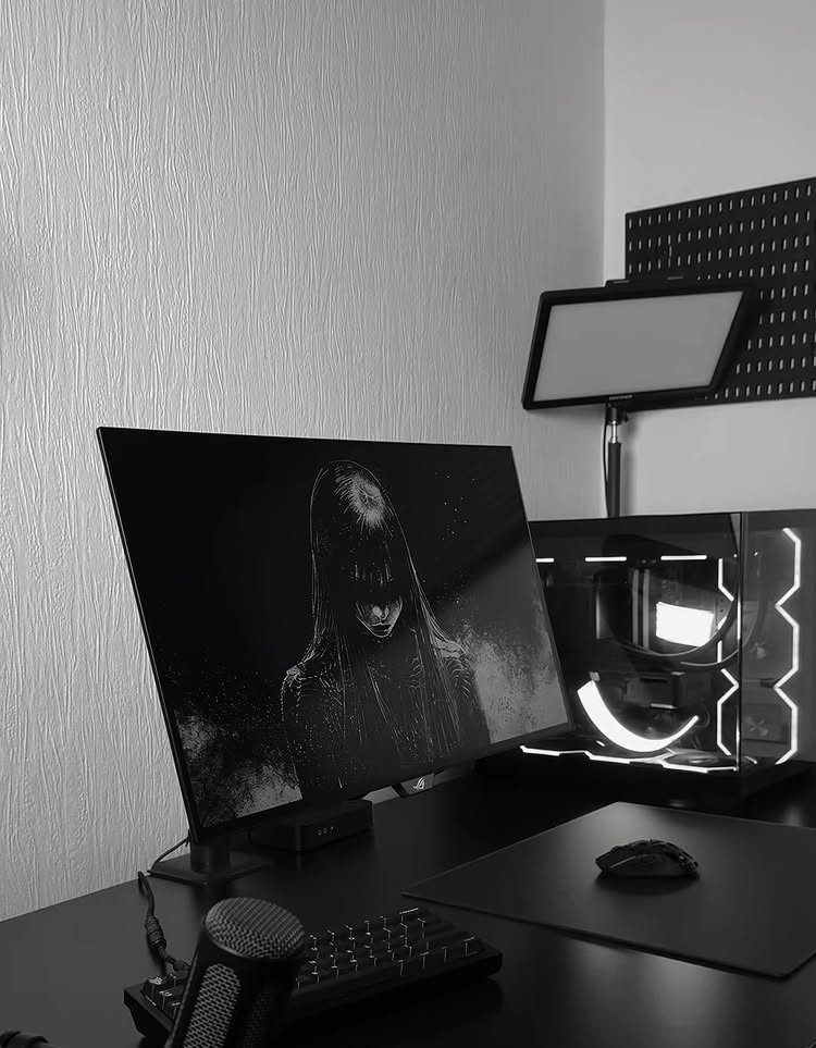 Black gaming desk setup with monitor, PC, and microphone against white wall.