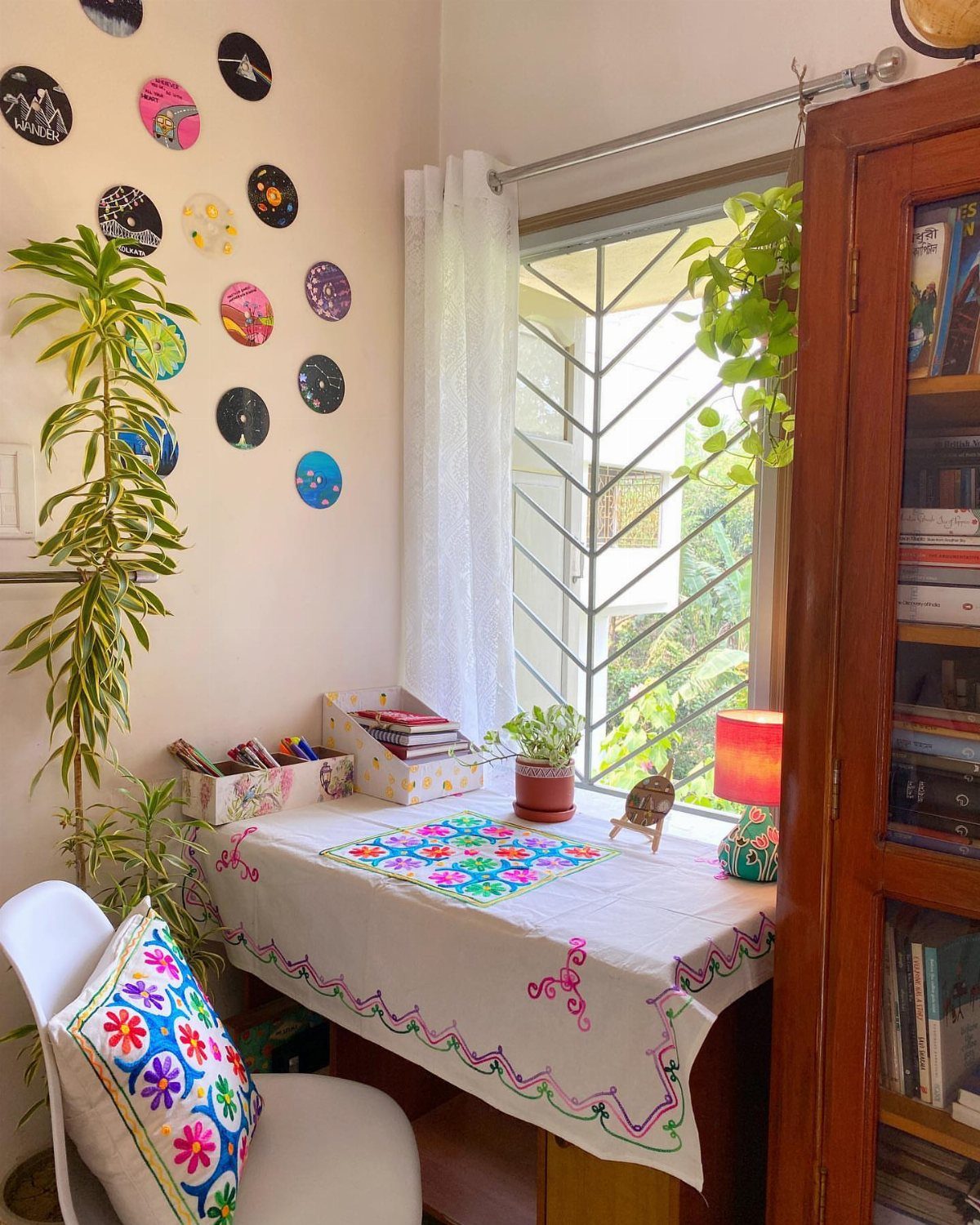 Desk with embroidered cloth placed under a window with a white chair.
