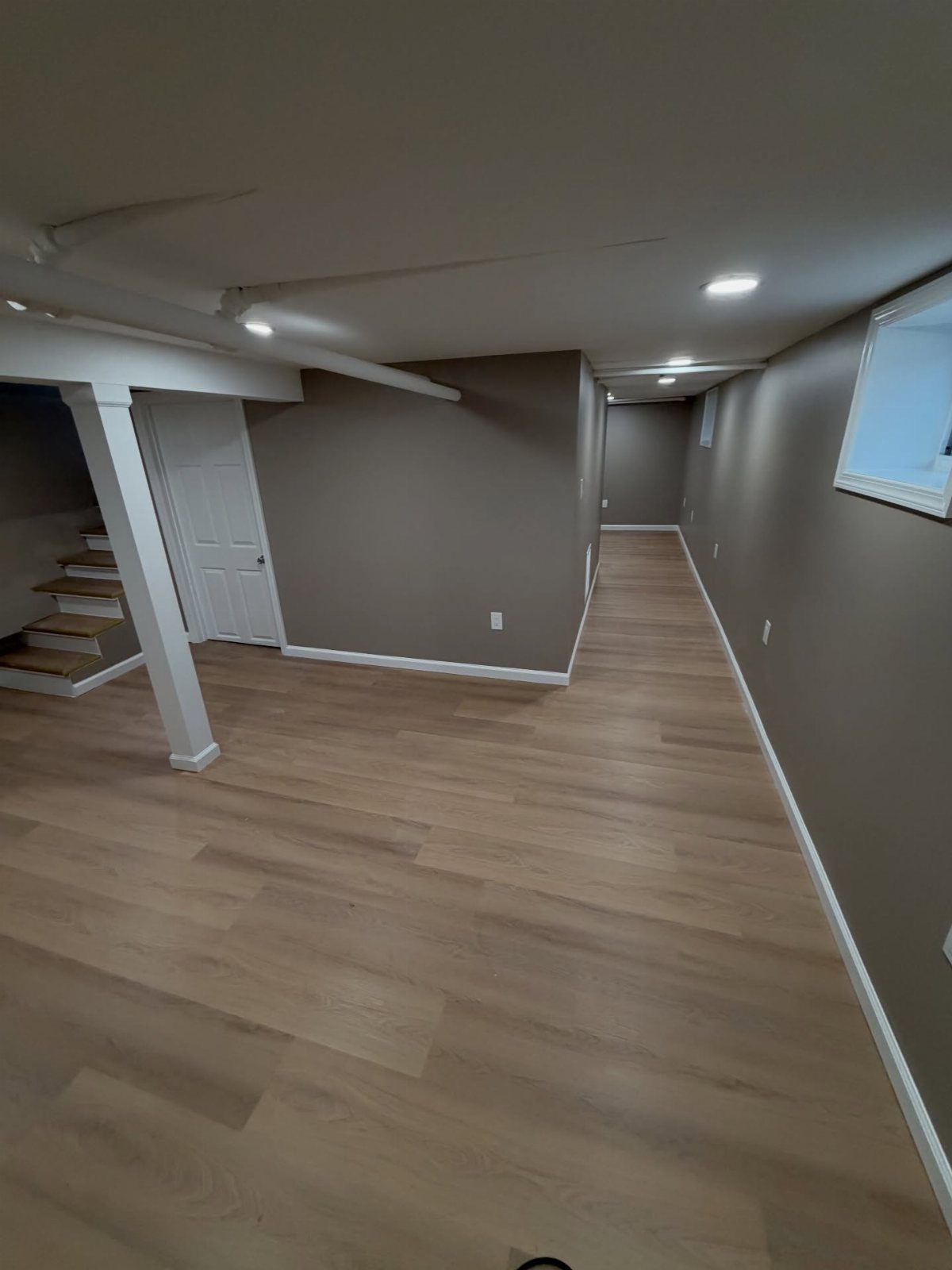 Finished basement with light wood floors, taupe walls, and white trim and pillars.