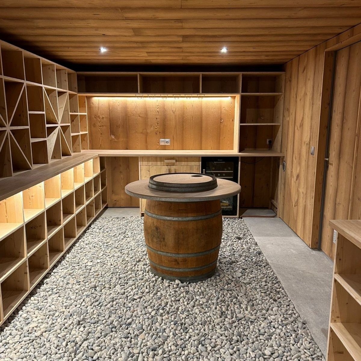 Wooden wine cellar with a barrel table and pebble flooring and built-in shelving.