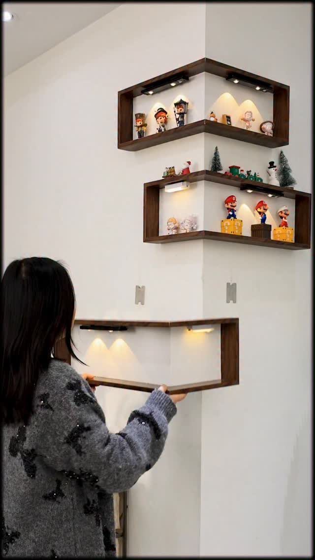 Woman installing brown wooden floating corner shelves displaying small video game figurines.