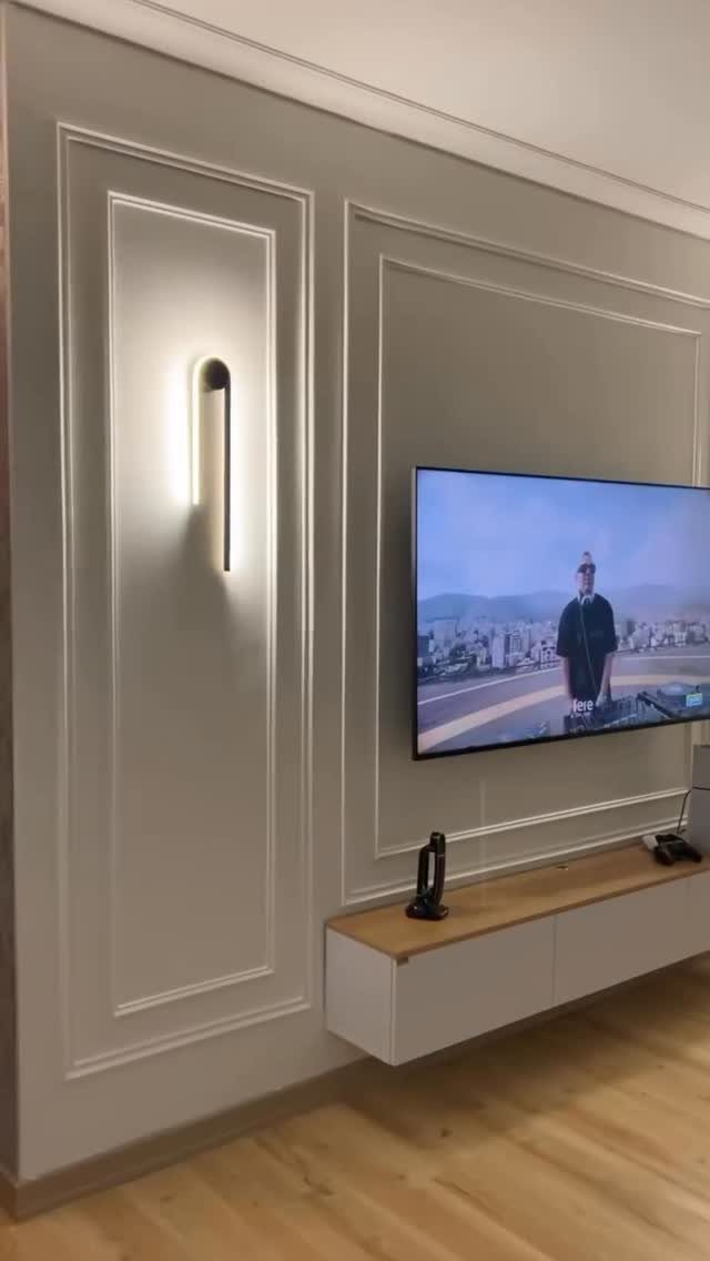 Vertical glowing wall sconce mounted on a grey paneled wall beside a television.
