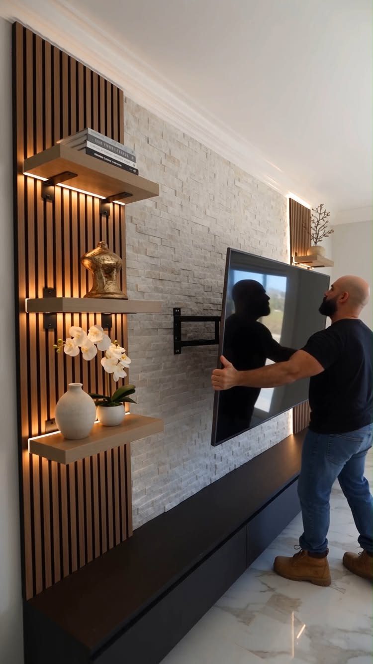Man mounting a television next to vertical wood paneling with floating shelves